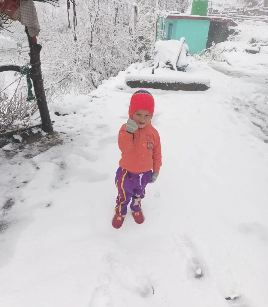 A child playing joyfully in the snow at Auli with snow-covered slopes and pine trees in the background.