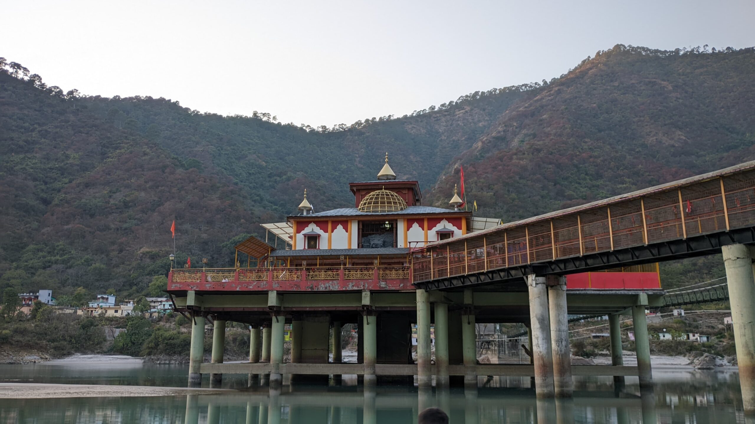 Riverside temple on pillars with footbridge and mountain backdrop in Uttarakhand