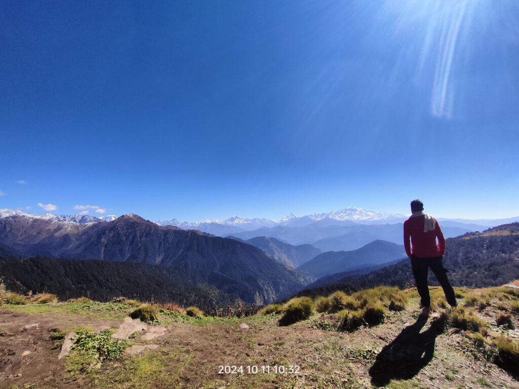 Person standing on a mountain ridge overlooking snow-capped Himalayan peaks under a clear blue sky in Uttarakhand